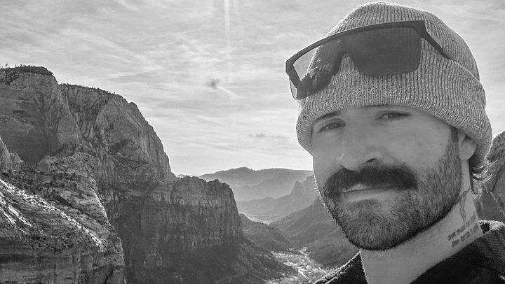 Chris O'Donnell mit Mütze und Sonnenbrille vor einer beeindruckenden Berglandschaft. Im Hintergrund erstrecken sich steile Felsen und ein tiefes Tal unter einem bewölkten Himmel.