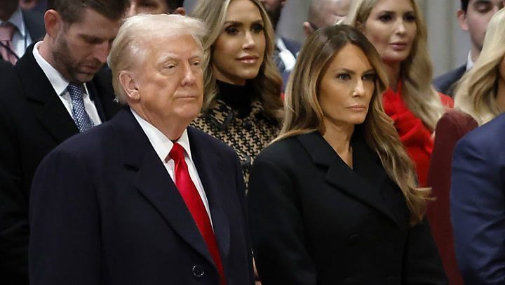 Donald und Melania Trump in formeller Kleidung bei einer öffentlichen Veranstaltung, umgeben von weiteren Gästen.