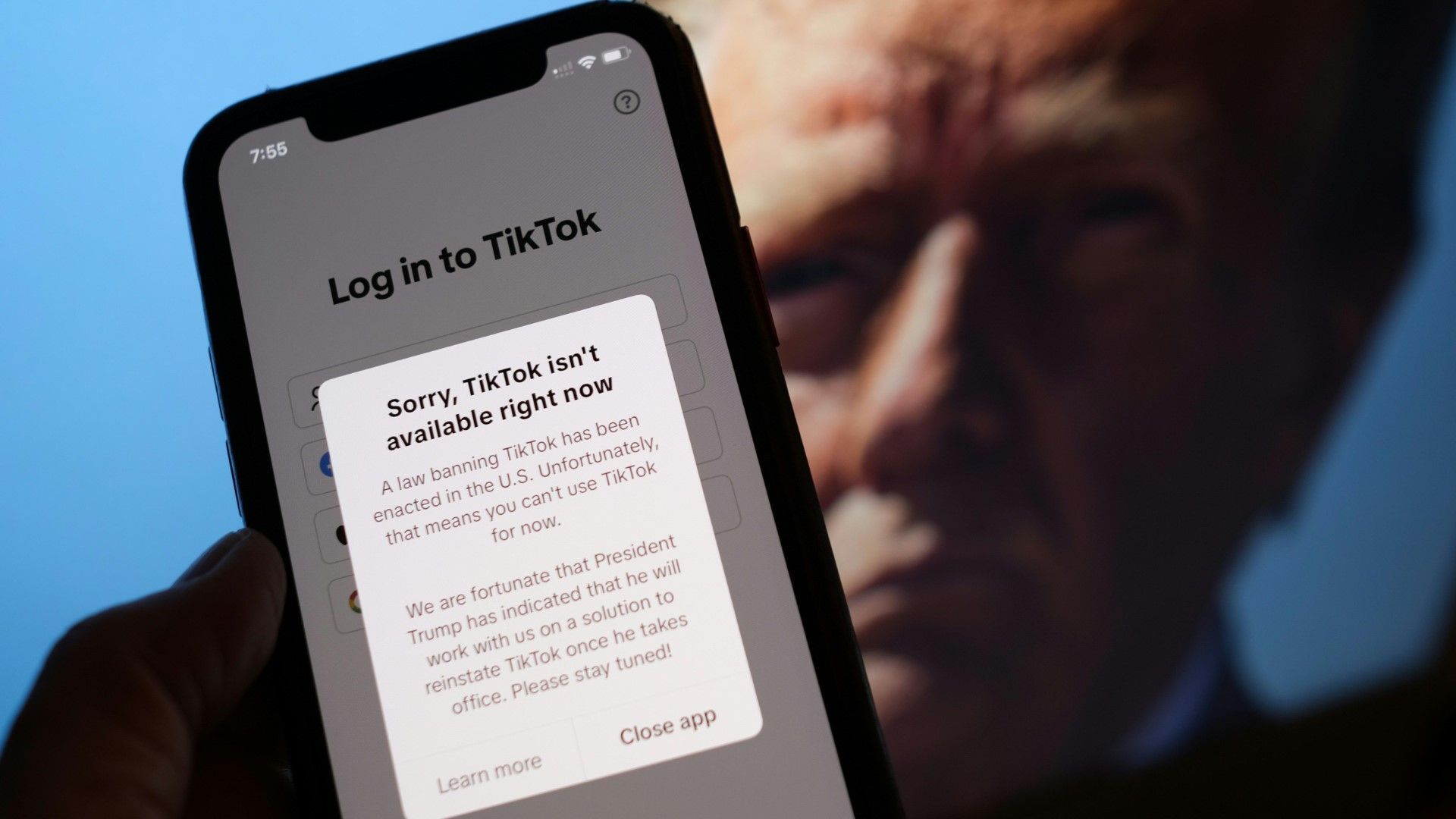 Ein Smartphone zeigt eine Fehlermeldung an, die besagt, dass TikTok aufgrund eines Gesetzes in den USA derzeit nicht verfügbar ist. Die Meldung erwähnt, dass eine Lösung in Arbeit ist. Im Hintergrund ist ein unscharfes Bild von Donald Trump zu sehen.