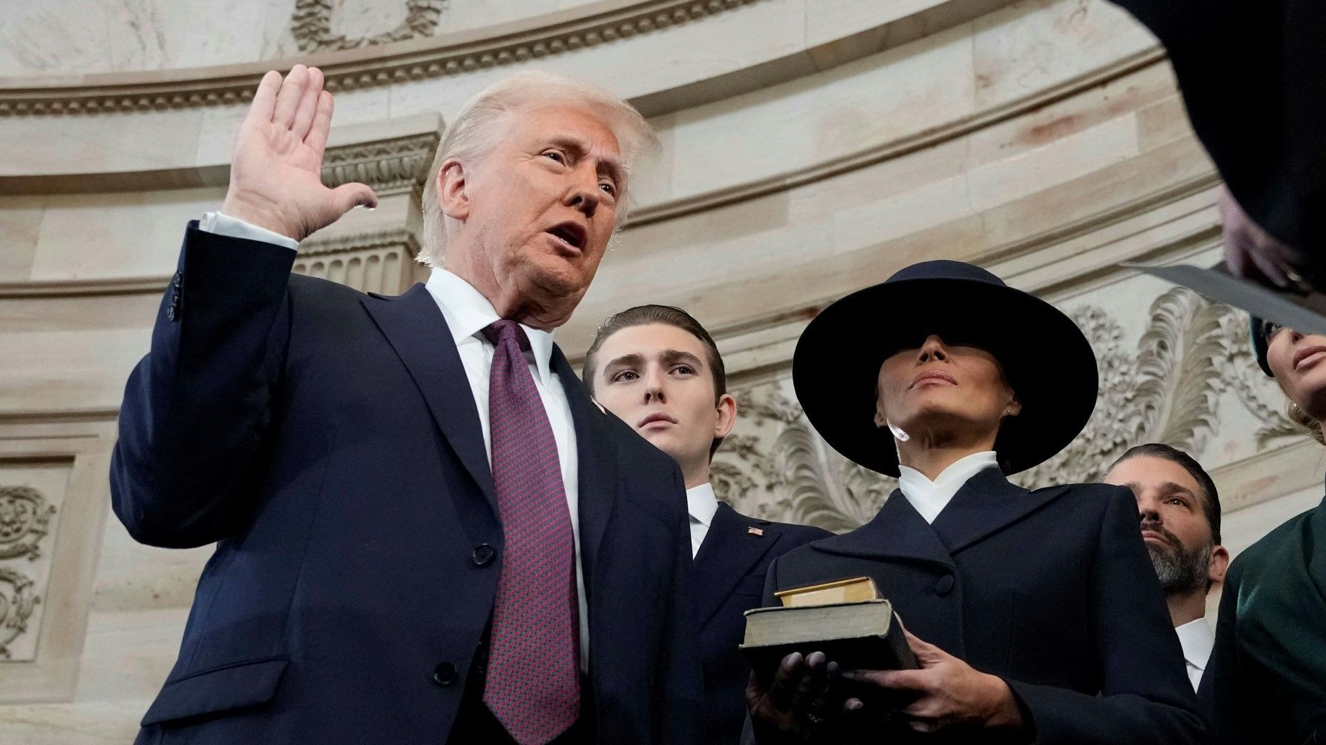 Donald Trump hebt seine rechte Hand und legt einen Eid ab, während eine Frau in dunkler Kleidung und Hut eine Bibel hält. Im Hintergrund sind mehrere Personen in einer prunkvollen Halle zu sehen.